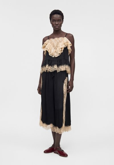 Γυναίκα που φοράει μαύρο αμάνικο φόρεμα με μπεζ δαντελένιες βολάν και επένδυση, σε συνδυασμό με σκούρα κόκκινα παπούτσια Mary Jane, στέκεται μπροστά από απλό φόντο.