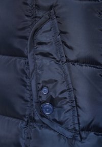 Bolsillo de chaqueta acolchada azul marino con dos botones a presión, con costuras visibles y ligeras arrugas en la tela.