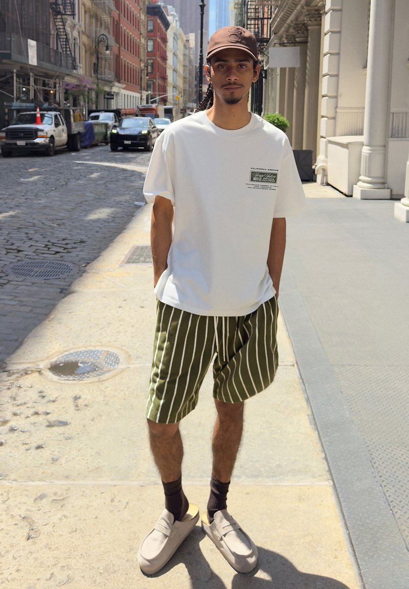 Homme aux cheveux tressés se tenant dans une rue de la ville, portant une casquette marron, un T-shirt blanc, un short rayé vert olive, des chaussettes noires et des chaussures beiges sans lacets.