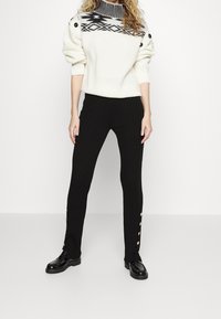 Vit stickad tröja med svarta geometriska mönster, hög krage, och svarta avsmalnande ribbade byxor med sidoknäppning. Svarta skor.