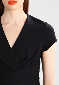 Czarna sukienka w stylu kopertowym z krótkimi rękawami, wykonana z miękkiego materiału, z głębokim dekoltem w kształcie V oraz marszczonym wykończeniem w talii, które zapewnia strukturę i dopasowanie.