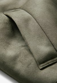 Tela de ante en verde oliva con una textura suave; presenta un solapa doblada que añade dimensión y un brillo sutil a la superficie.
