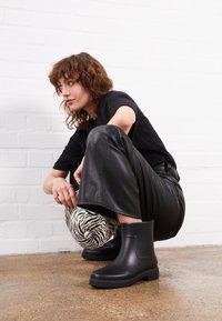 Las botas negras hasta el tobillo tienen un acabado mate suave, punta redondeada y tacón cuadrado. La modelo lleva pantalones de cuero negros y una blusa negra, en cuclillas.