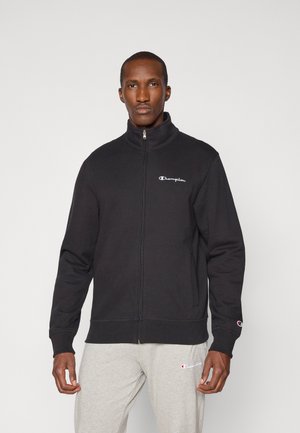 Homme portant une veste noire zippée et un pantalon gris clair, tous deux avec des logos Champion, debout devant un fond clair uni.