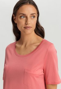 T-shirt z krótkim rękawem i okrągłym dekoltem w kolorze koralowym, z małą kieszonką na piersi i gładką teksturą, prezentowana przez kobietę o falowanych włosach.
