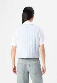 Lys blå quiltet vest med krage og ermeløst design, med vertikale sømmer og en cropped hem, kombinert med en hvit skjorte.