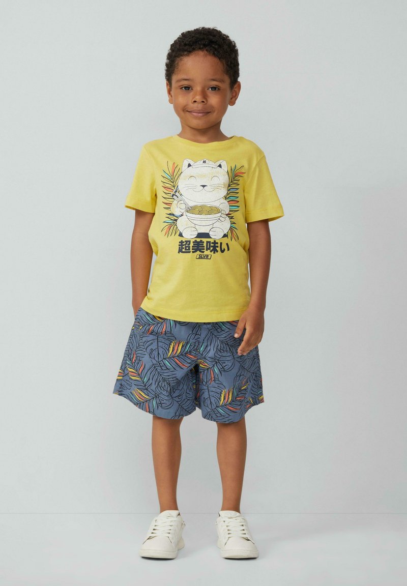 Gelbes Baumwoll-T-Shirt mit einem Cartoon-Katzen- und Nudel-Graphic, kombiniert mit blauen Shorts, die bunte Blattmuster zeigen. Weiße Sneaker.