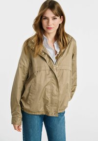 Jasnobeżowa kurtka przeciwwiatrowa z zapięciem na zamek, kapturem, bocznymi kieszeniami i elastycznymi mankietami, noszona na jasnoniebieskiej koszuli z kołnierzykiem.