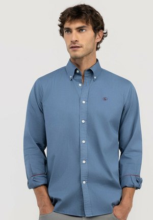 Hombre con una camisa azul abotonada, con las mangas remangadas, de pie con las manos en los bolsillos, mirando ligeramente hacia la derecha.