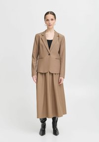 Barna szabott zakó, két elülső zsebbel, egy hozzá illő hosszú szoknyára rétegezve. Elegáns fekete csizmák teszik teljessé az öltözéket. Minimalista dizájn.