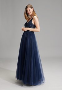 Giovane donna con capelli lisci che indossa un abito da sera senza maniche di colore blu navy decorato con perline, in piedi contro uno sfondo grigio uniforme.