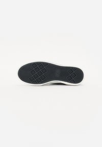 Svart gummisåle til sneakers med et teksturert fiskebensmønster og hvit kant. Logoen vises i midten.