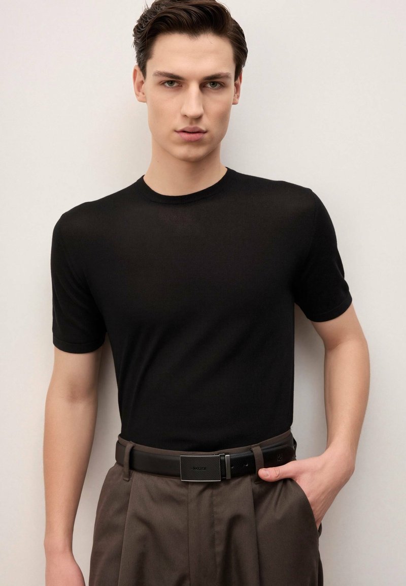 Młody mężczyzna z krótkimi ciemnymi włosami, ubrany w dopasowaną czarną koszulkę z krótkim rękawem, brązowe spodnie plisowane i czarny pasek, stojący na jednolitym tle.