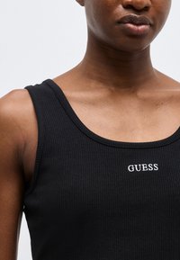 Εγγύς λήψη ατόμου που φοράει μαύρο ριμπ μπλουζάκι χωρίς μανίκια με το λευκό λογότυπο "GUESS" κεντημένο στο στήθος, πάνω σε απλό φόντο.