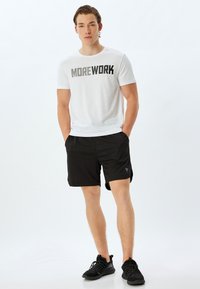 Hvid T-shirt med teksten "MOREWORK" i sort, parret med sorte sportsshorts. Model iført sorte sneakers står afslappet med hænderne i lommerne.