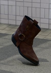 Καφέ δερμάτινη μποτάκια αστραγάλου με διακοσμητική αγκράφα και ραφές, τοποθετημένη σε υφή εδάφους με φόντο πλακιδωτούς τοίχους.