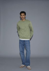 Giovane uomo in piedi con le mani dietro la schiena, indossa un felpa verde chiaro, jeans blu e scarpe da ginnastica grigio-bianche, su uno sfondo grigio uniforme.