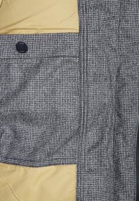 Grau und schwarzes Hahnentritt-Muster mit einem einfarbigen beigen Futter; verfügt über einen Knopf und Taschen, die ein strukturiertes Design zeigen.