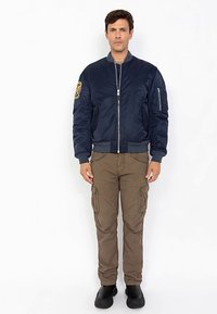 Man in een marineblauwe bomberjack met een patch op de mouw, bruine cargo-broek en zwarte schoenen, staand tegen een witte achtergrond.