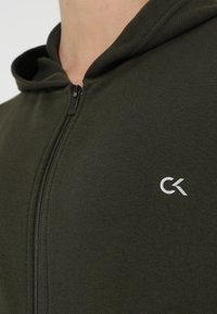 Gros plan sur une personne portant un sweat à capuche zippé vert foncé avec un petit logo CK blanc sur la poitrine.