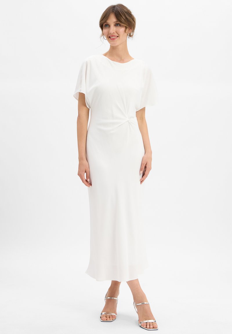 Femme portant une robe midi blanche à manches courtes et détail de torsion sur le côté, associée à des sandales à talons hauts argentées, se tenant devant un fond uni.