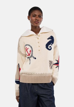 Femme portant un pull beige en maille avec des motifs nautiques comprenant une ancre, un hippocampe et une boussole, assorti à un pantalon foncé.
