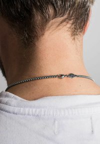 Colier din argint cu un design texturat, dotat cu un închizător și o piesă decorativă. Purtați în jurul gâtului, parțial vizibil sub o cămașă albă.