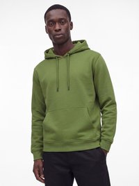 Man draagt een groene capuchontrui met voorkantzakje en trekkoorden, poserend tegen een effen witte achtergrond.