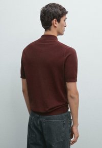 Ung mand står ryggen til iført en kortærmet bordeaux polo-shirt og sorte denimjeans mod en ensfarvet lys grå baggrund.