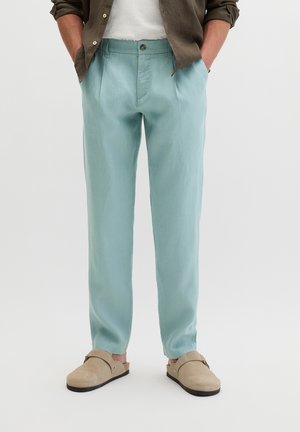 Mężczyzna ubrany w jasnoniebieskie spodnie o prostych nogawkach, beżowe buty wsuwane, białą koszulę i brązową otwartą koszulę na guziki, ręce w kieszeniach.