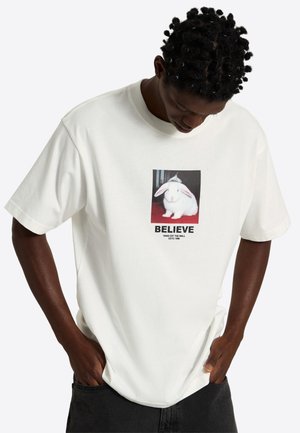 Mand iført en hvid T-shirt med et foto af en hvid kanin og ordet "TRO" trykt under billedet.