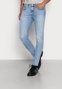 Hombre vistiendo jeans ajustados azul claro y zapatos de cuero negro con cordones, de pie sobre un suelo gris claro contra una pared blanca.
