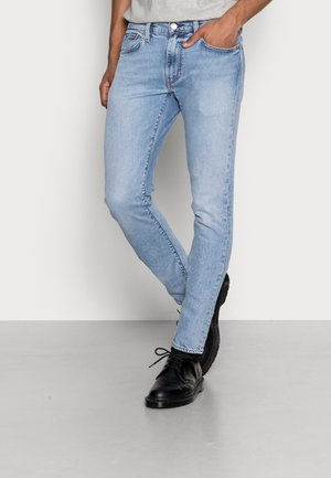 Homme portant un jean slim bleu clair et des chaussures noires en cuir à lacets se tenant sur un sol gris clair contre un mur blanc.