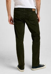 Pantaloni in velluto a coste verdi con una vestibilità slim, dotati di tasche posteriori e un patch in pelle con logo in vita. Abbinati a sneaker in tela scura.