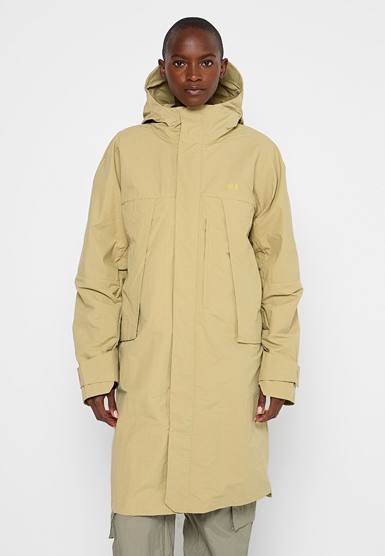 Persona vestida con un impermeable largo beige con capucha, que tiene bolsillos delanteros y puños ajustables, de pie contra un fondo blanco plano.