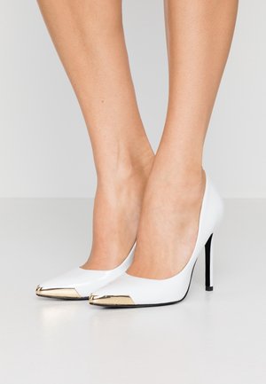 Białe szpilki z błyszczącym wykończeniem, w szpic, z złotym akcentem na nosku. Czarne podeszwy i elegancki design.