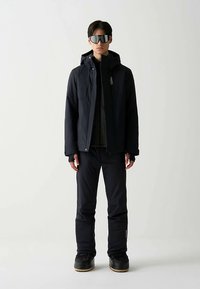 Zwarte winterjas met capuchon, gedragen over een donker shirt en bijpassende broek. Model draagt reflecterende zonnebril en zwarte schoenen. Gladde stoftextuur.