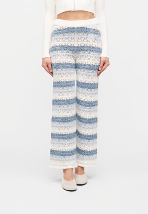 Γυναίκα που φοράει πλεκτό παντελόνι με ψηλή μέση και φαρδιά μπατζάκια με οριζόντιες μπλε και λευκές ρίγες, συνδυασμένο με λευκό κοντό πουλόβερ και μπεζ παπούτσια.