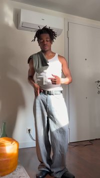 Νέος άνδρας με λευκό αμάνικο και φαρδιά τζιν πόζάρει σε εσωτερικό χώρο, με γκρι πουκάμισο περασμένο πάνω από τον έναν ώμο και κλειστά μάτια.