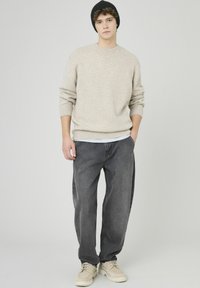 Pull en maille beige avec poignets côtelés, associé à un jean ample couleur charbon et des baskets claires. Le modèle porte un bonnet noir.