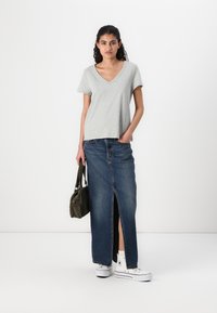 Szara koszulka typu v-neck, dżinsowa spódnica maxi z rozcięciem z przodu i białe trampki. Modelka trzyma małą oliwkowo-zieloną torbę. Prosty, casualowy strój.