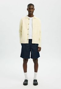 Krämf ärgad skjorta med knappar och krage över en vit skjorta, tillsammans med mörka denimshorts. Svarta skor och vita strumpor fulländar looken.