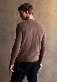 Brauner, strukturierter Pullover mit Rundhalsausschnitt, figurbetontem Design und langen Ärmeln, kombiniert mit dunklen Denim-Jeans, von hinten betrachtet.