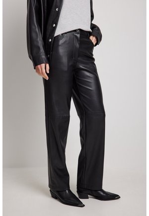 Personne portant un pantalon en cuir noir, une veste en cuir noire, un haut gris et des bottes noires à bout pointu, debout sur un sol clair avec un fond uni.