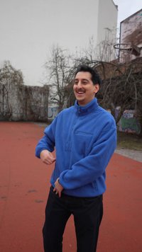Mann in blauer Fleecejacke lächelt und gestikuliert mit den Händen auf einem Basketballplatz im Freien mit kahlen Bäumen und Graffiti-Wänden.
