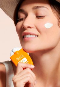 Avene sunscreen in an orange bottle with white cap, held near a cheek with white cream applied. Background is soft pink.