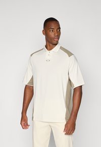 Polo à manches courtes beige avec des accents vert olive sur les épaules, fabriqué en tissu respirant, avec un col et un détail de logo.