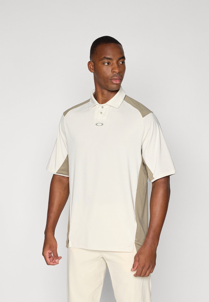 Polo à manches courtes beige avec des accents vert olive sur les épaules, fabriqué en tissu respirant, avec un col et un détail de logo.