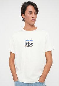 Młody mężczyzna w białej koszulce z małym, vintage'owym motywem załogi Pan Am oraz niebieskich dżinsach, z rękami w kieszeniach, spoglądający na bok.