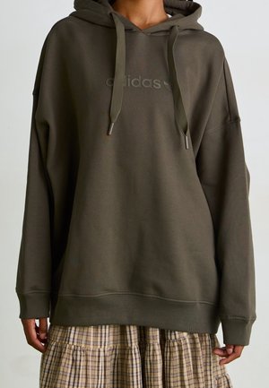 Brown oversized Adidas hoodie with drawstrings worn over a beige and brown plaid skirt on a person with brown skin.